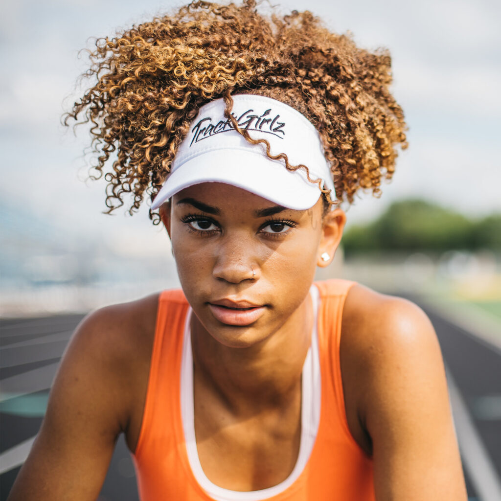 Track and field girl wearing a white TrackGirlz visor and an orange tank top shirt, staring at the camera/viewer
